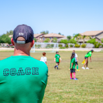 A coach in a green t-shirt with "coach" written on the back is facing the girls soccer team he is coaching.