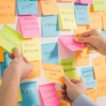 Colorful sticky notes with creative buzz words are attached to a white board with hands reaching out towards them.